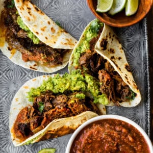 a photo of several shredded beef tacos sitting on baking sheet with a bowl of salsa sitting next to them and several lime wedges