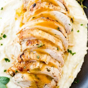 a photo of sliced turkey breast drizzled with gravy and garnished with fresh herbs on a bed of mashed potatoes all on a black plate