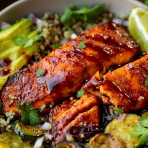 a photo of two salmon fillets glazed with bbq sauce sitting on a plate surrounded by grilled zucchini slices, lime wedges and sliced avocado