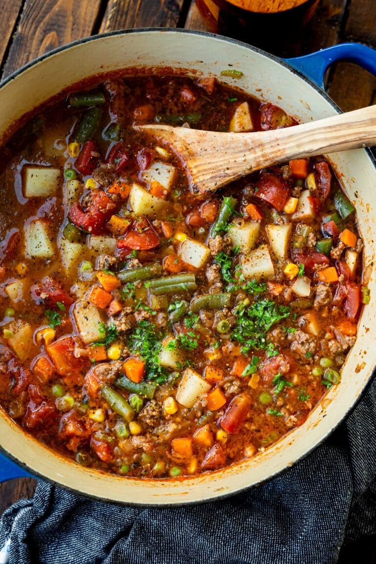 a large bot of ground beef vegetable soup topped with minced fresh parsley with a wooden spoon in the bowl