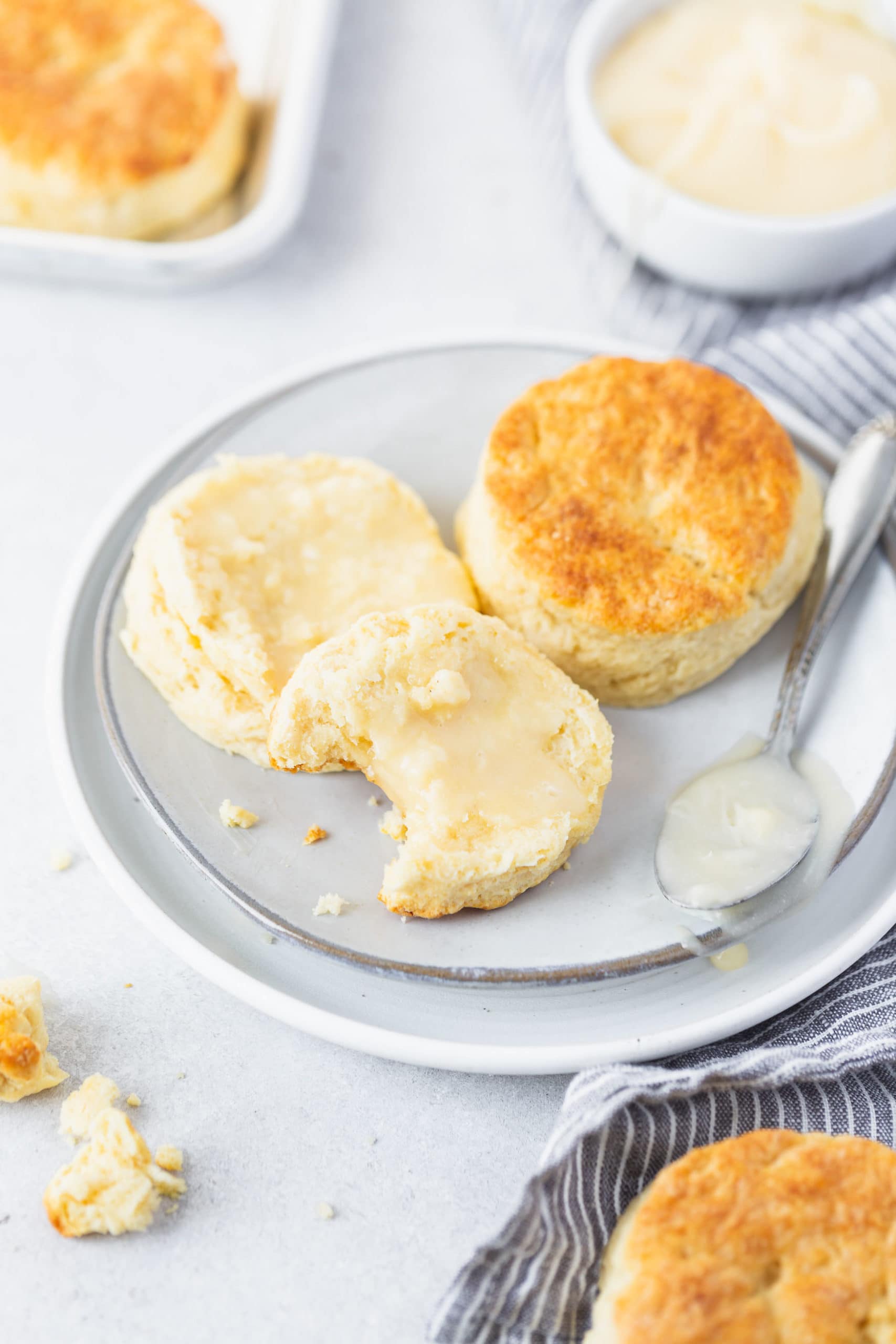 Buttermilk Biscuits Two buttermilk biscuits on a white plate. One biscuit is cut in half and spread with honey butter. A bite has been taken out of one biscuit half.