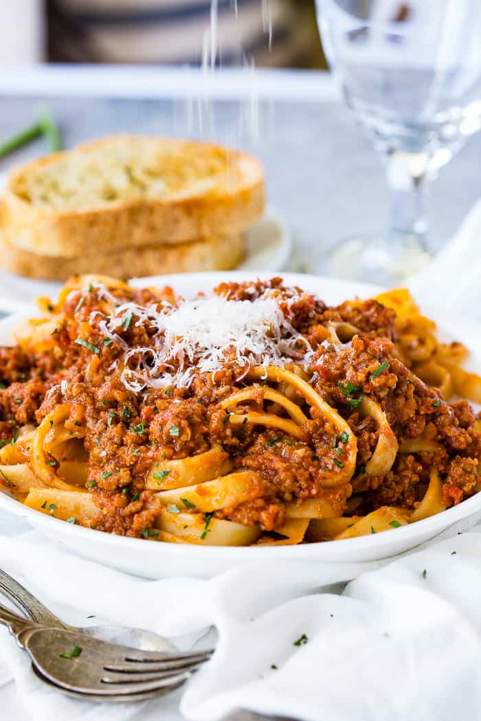 A white pasta dish full of fettuccini loaded with authentic Italian Bolognese sauce and fresh parmesan cheese on top with garlic bread in the background