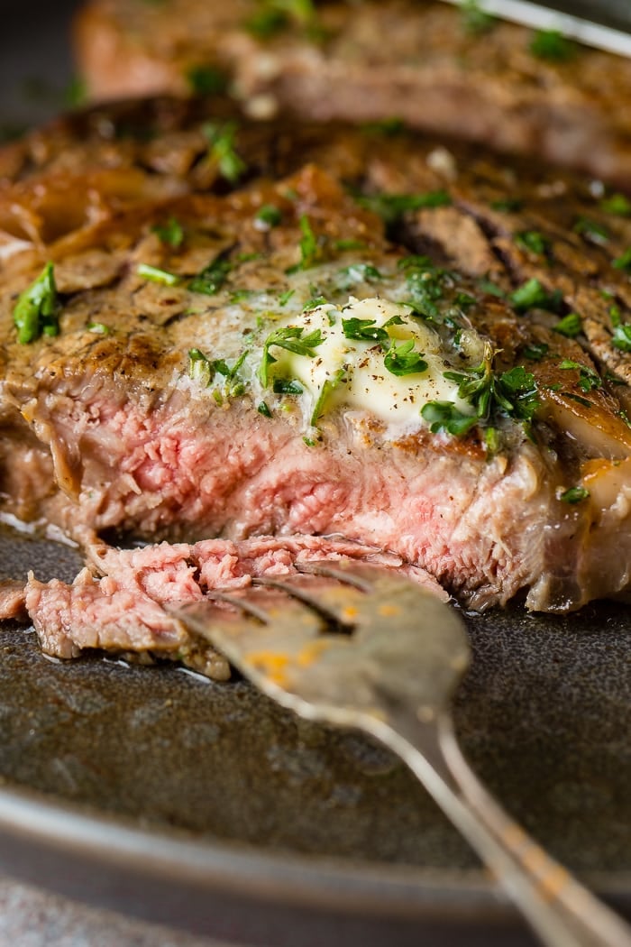 a grey plate with a medium rare garlic butter steak being cut into and a fork taking a piece with steak butter melting on top with parsley