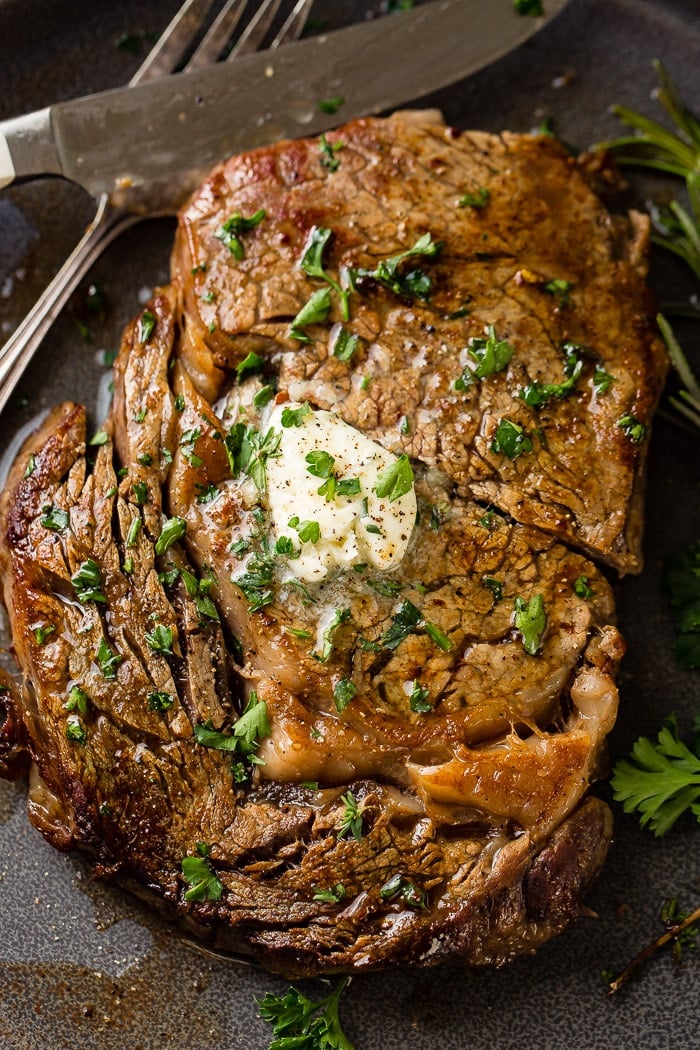 a grey plate with a medium rare ribeye steak being cut into and a fork taking a piece with garlic butter melting on top with parsley