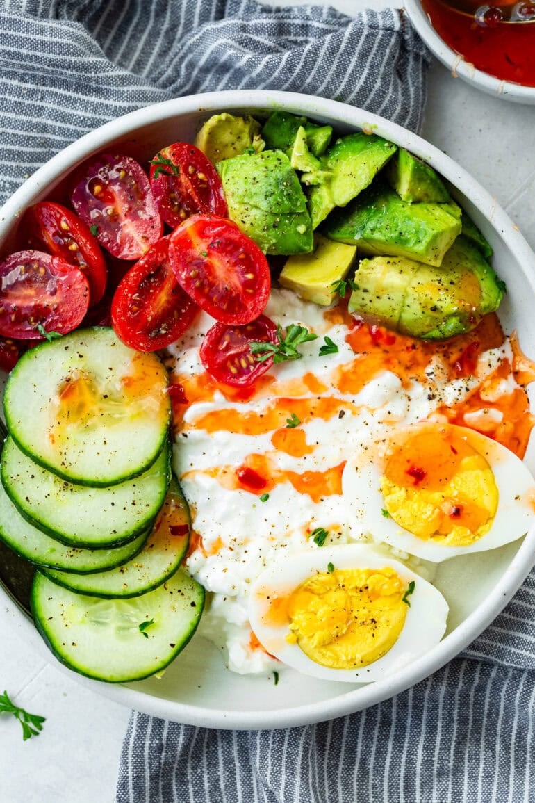 a photo of a bowl full of cottage cheese topped with hard boiled eggs, cucumber slices, avocado chunks, cherry tomatoes and sweet chili sauce