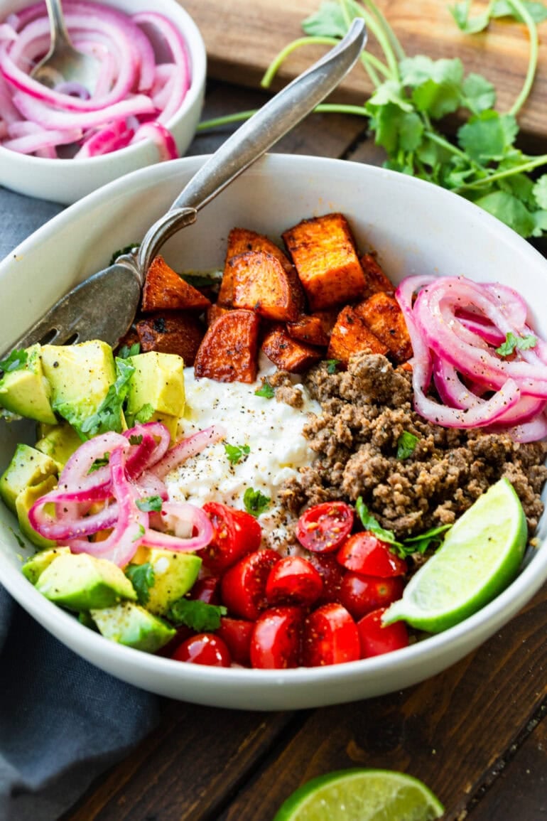 a photo of a ground beef and sweet potato bowl topped with avocado, pickled red onions, cottage cheese and cherry tomatoes