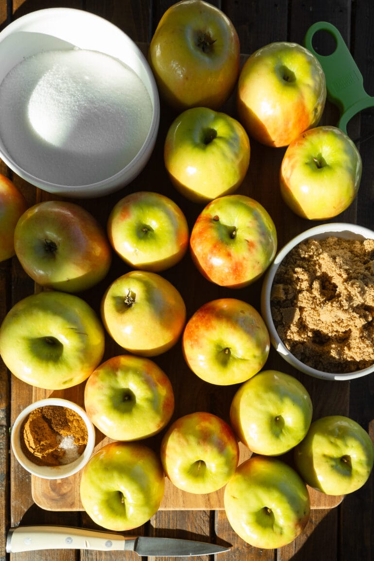 a photo of several whole unpeeled apples sitting on a cutter board with a cup of cinnamon sitting next to them and a bowl of brown sugar and a bowl of white sugar.