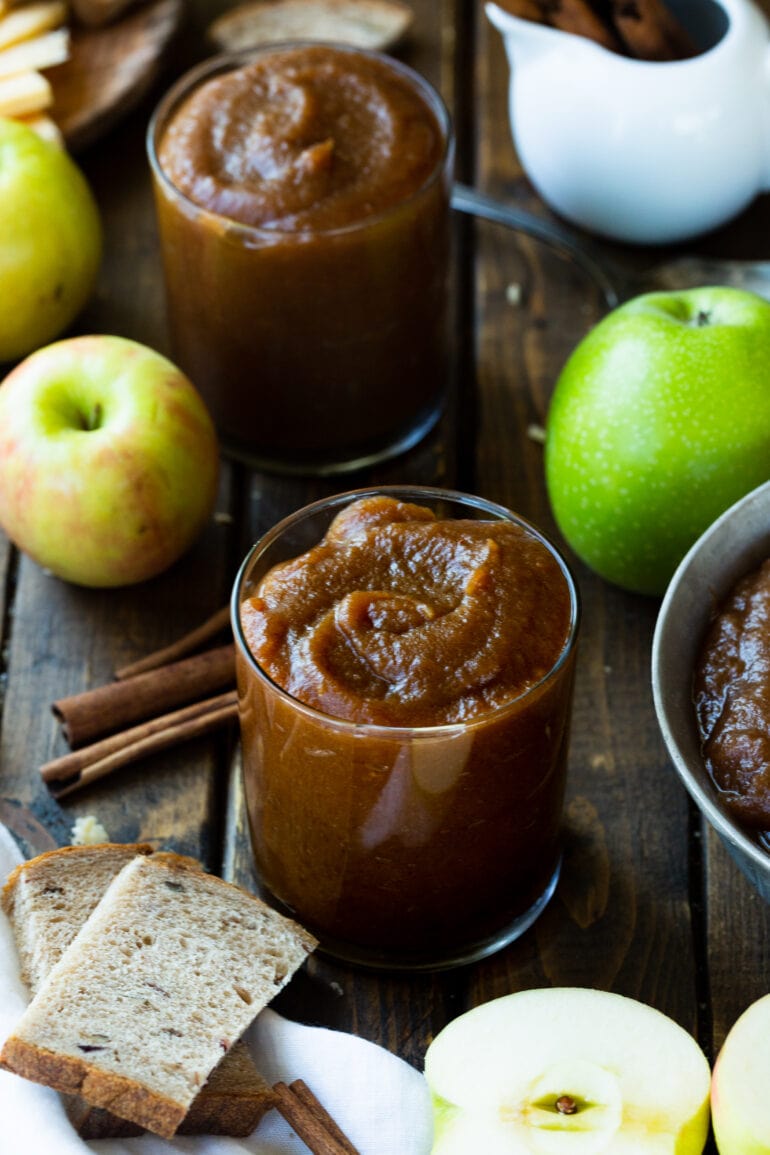 a photo of two glasses full of apple butter with cinnamon stick, whole apples, and peeled apples all around them.