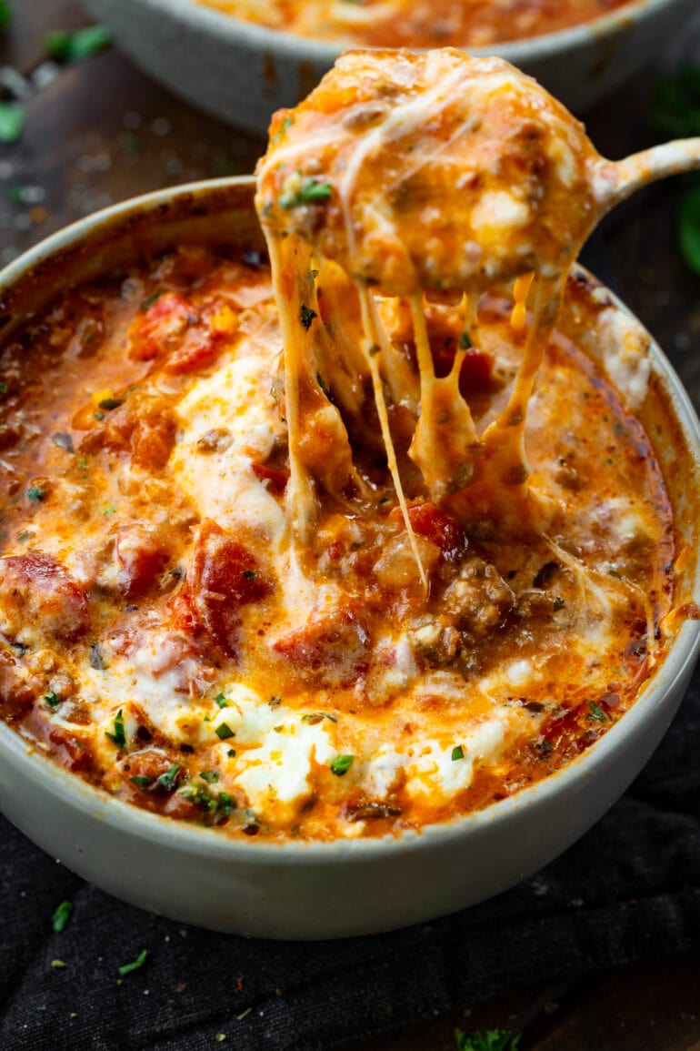 a photo of a bowl of lasagna soup topped with melted cheese and someone pulling a spoon out of the bowl with cheese stringing from it.