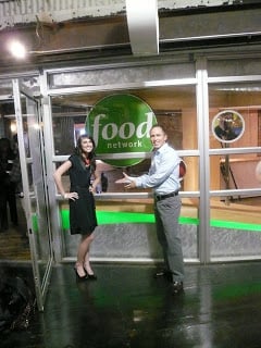 A photo of Carrian and her husband in front of the Food Network Logo at the Food Network Offices in New York.