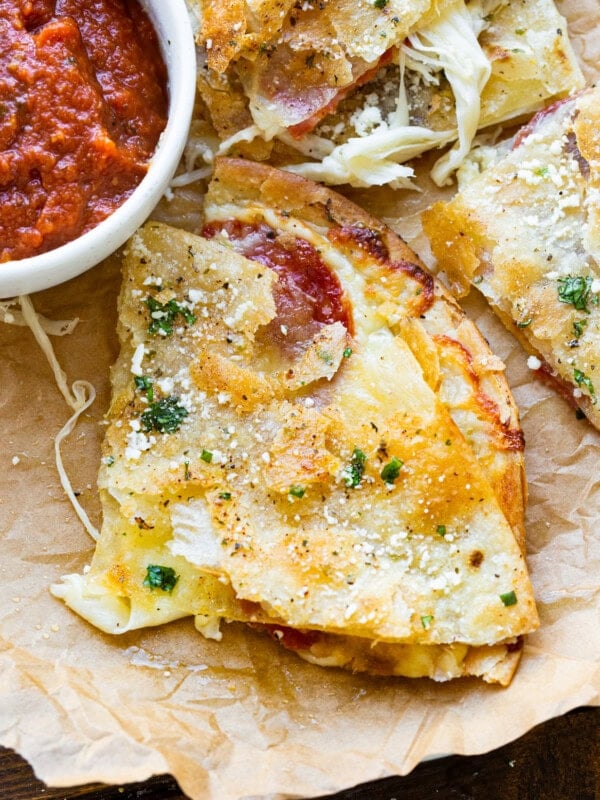 a photo of a slice of a tortilla pizza that is golden and crispy with melty cheese coming out and topped with sprinkled cotija cheese and garlic butter with a small bowl of pizza sauce next to it