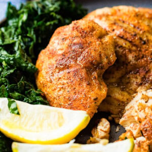 A photo of whole smoked chicken breasts and on a charcoal gray plate with lemon wedges and kale on the side.