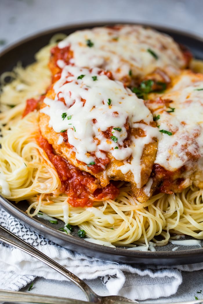A dark grey platter filled with angel hair pasta, a little marinara tomato sauce, breaded chicken with marinara and melted mozzarella cheese and a sprinkle of chopped Italian flat leaf parsley. Quick and Easy shortcut chicken parmesan