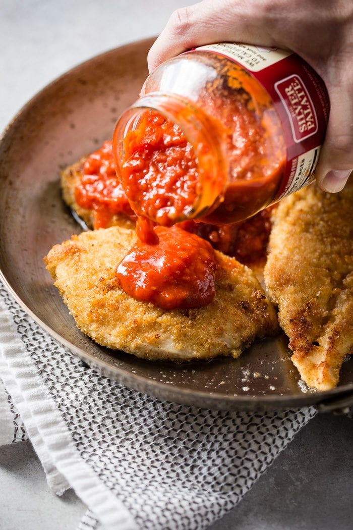 Golden brown breaded chicken filets in a skillet with a jar of marinara sauce being poured over each one.