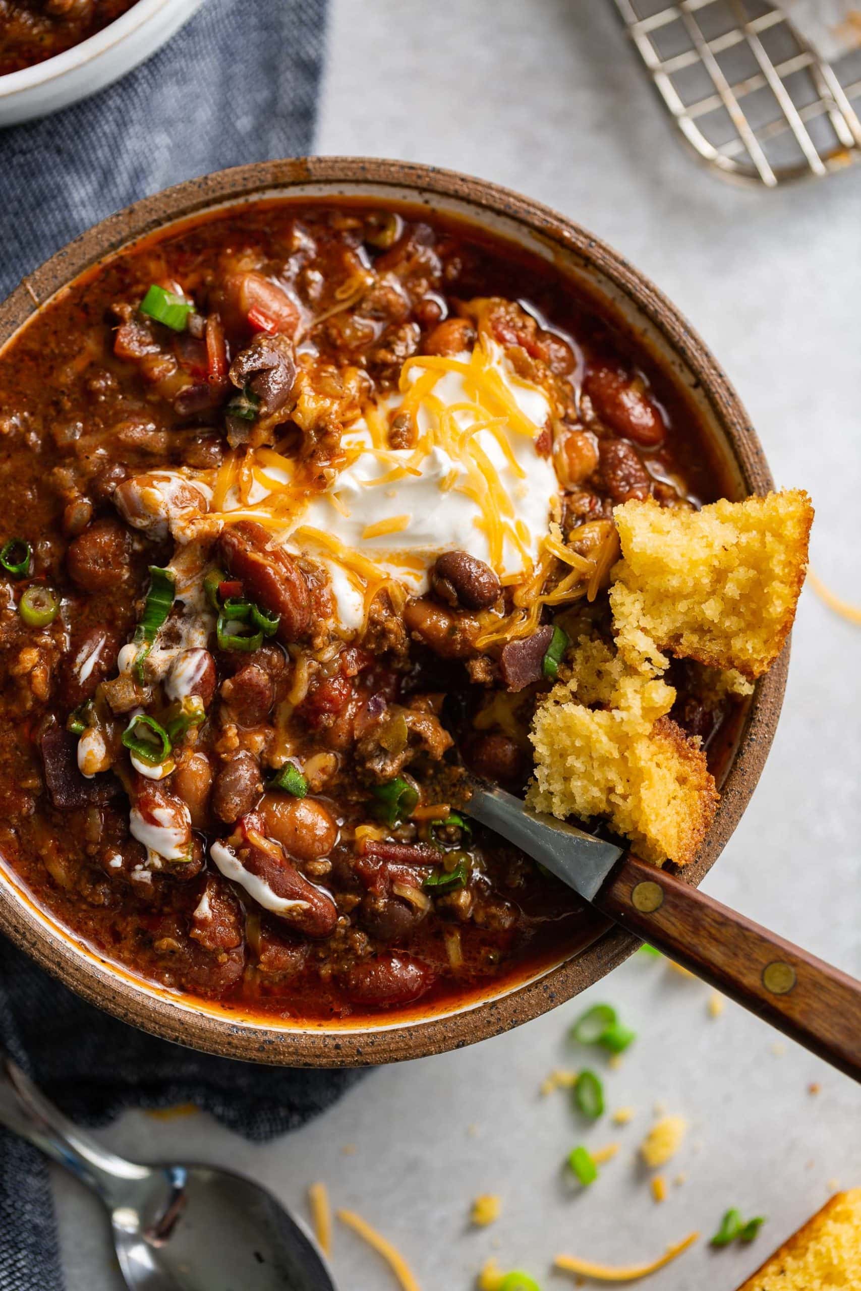 Best Chili Recipe A bowl of chili. The chili has ground beef, beans and a rich sauce. It is topped with sour cream and shredded cheese and a piece of cornbread is on the edge of the bowl.