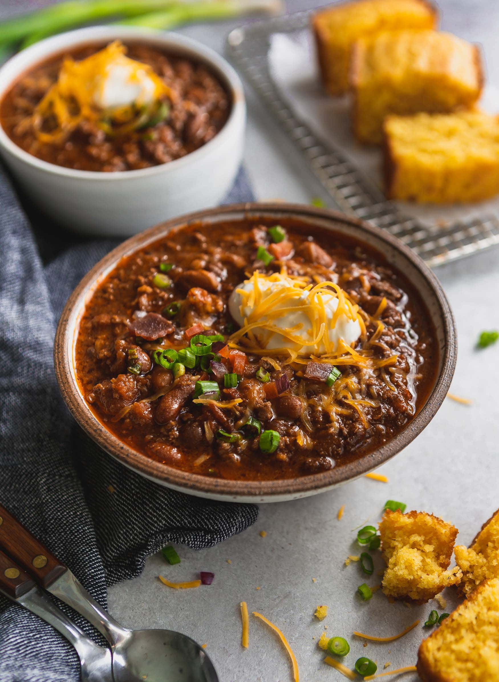 Best Chili Recipe A large bowl of the best chili topped with sour cream and grated cheese. Another bowl for chili is in the background and there are a few pieces of cornbread next to the chili.