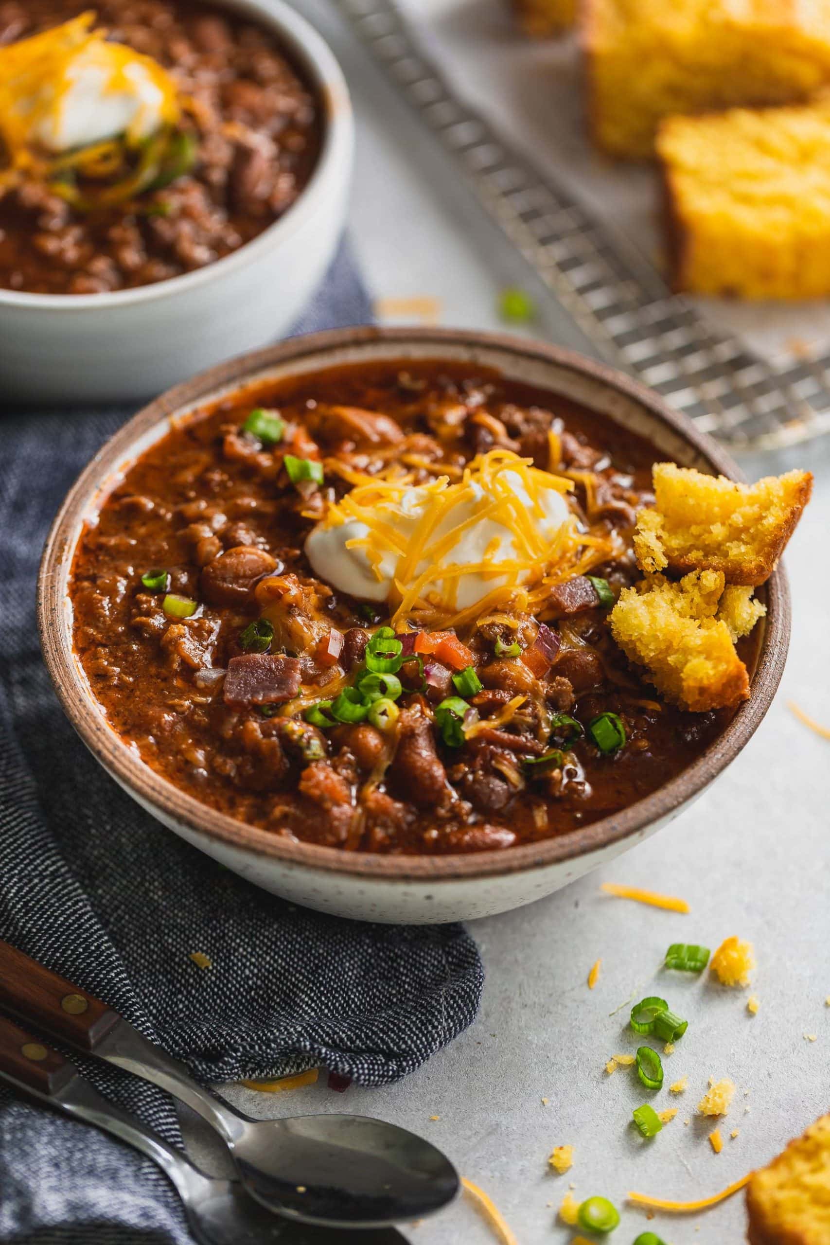 Best Chili Recipe A bowl of the best chili. It has a spoonful of sour cream and shredded cheese on top. You can see the ground beef and beans in the chili, and a piece of cornbread is on the edge of the bowl. Another bowl of chili is in the background.