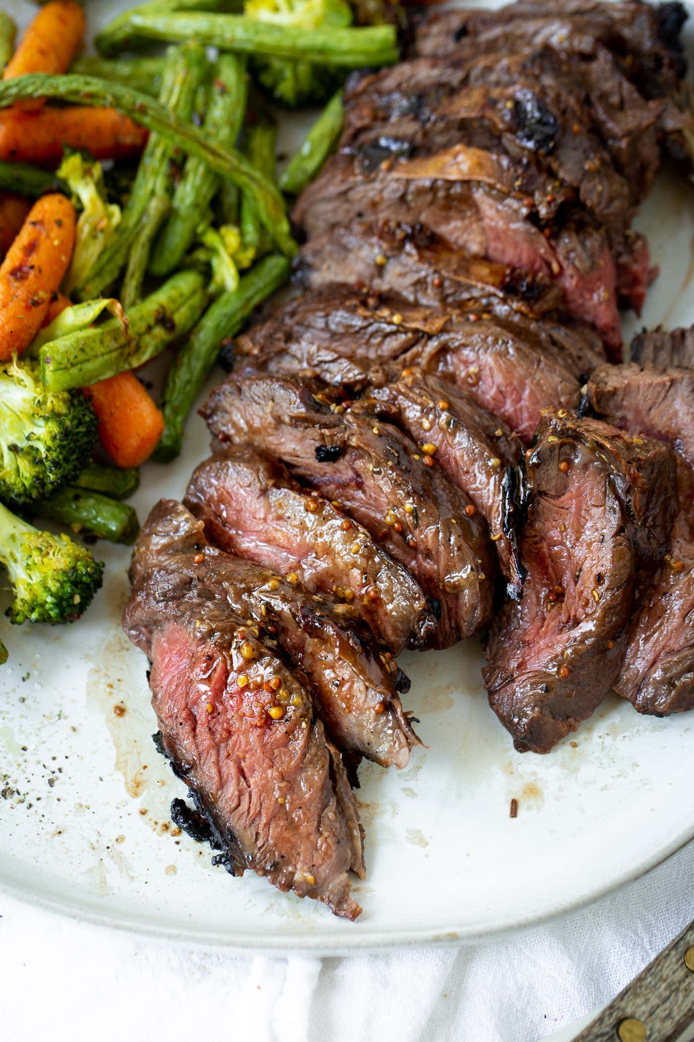 The Best Steak Marinade A photo of sliced medium rare hanger steak on a white plate next to a pile of roasted broccoli, baby carrots and green beans.