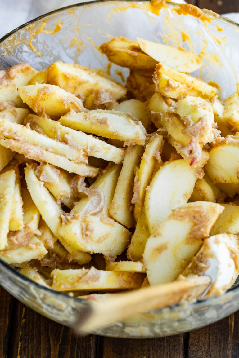a photo of a large glass bowl full of peeled apple slices coated in a creamy sugar and cinnamon mixture ready to be baked.