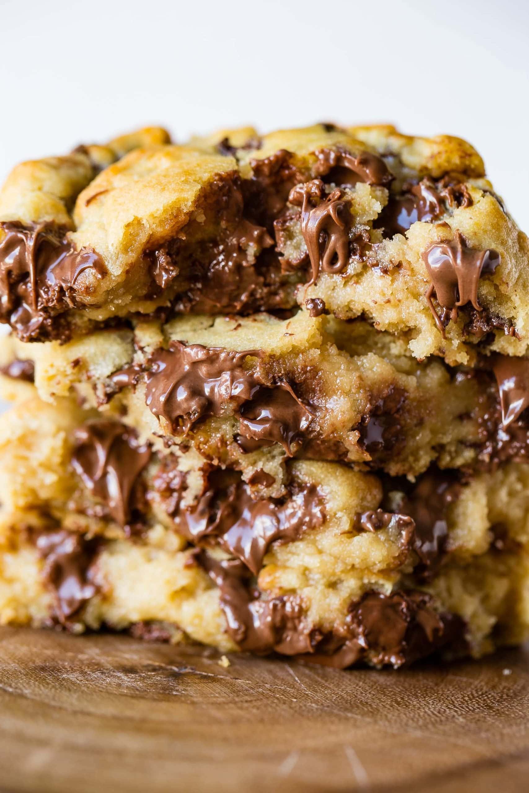 Levain Bakery Style Thick Chocolate Chip Cookies Recipe a closeup photo of four thick melty chocolate chip cookies that have been broken in half stacked on top of each other on top of a wooden plate.