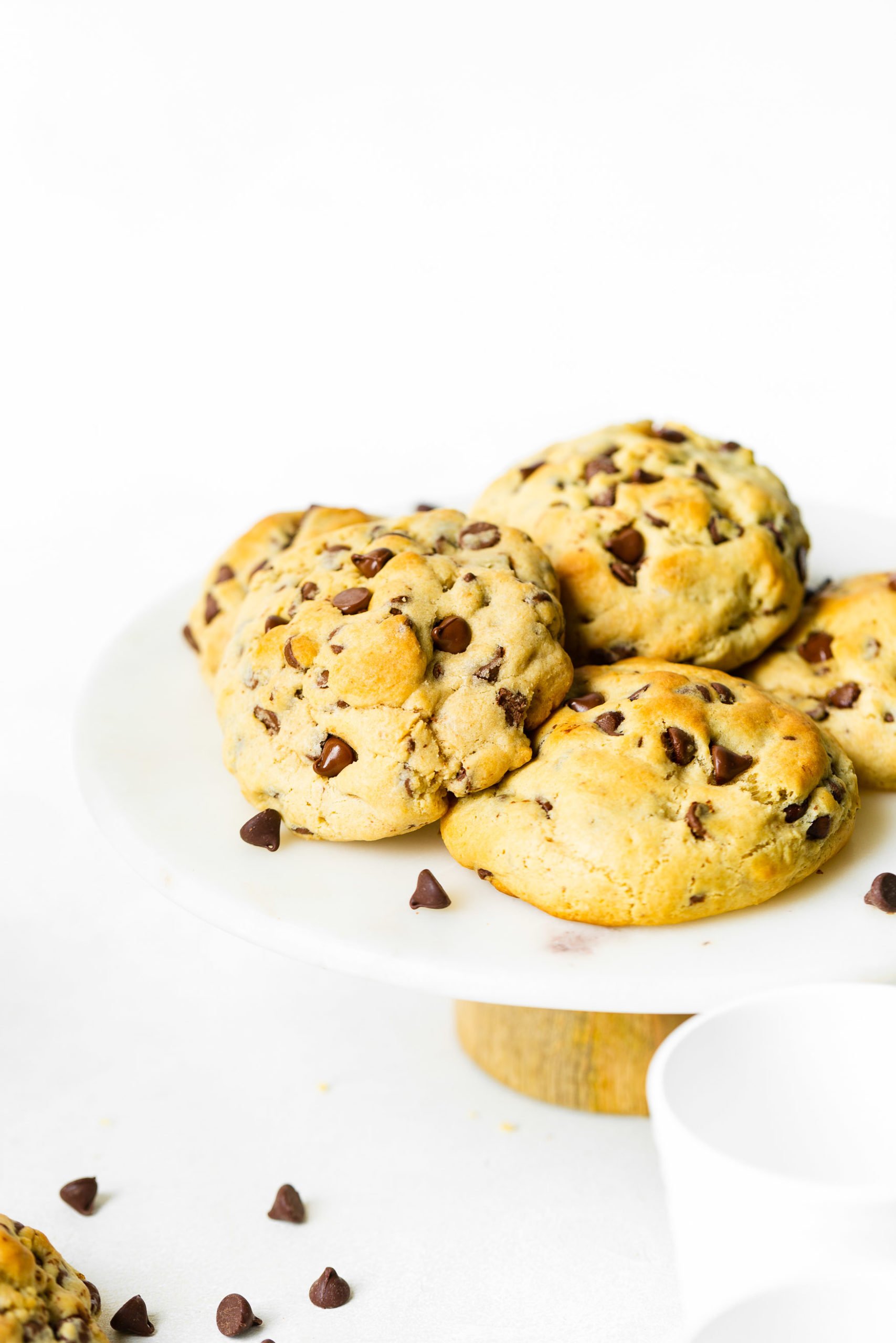 Levain Bakery Style Thick Chocolate Chip Cookies Recipe a photo of a pile of thick, perfectly golden chocolate chip cookies sitting on a white cake stand with chocolate chips scattered around.