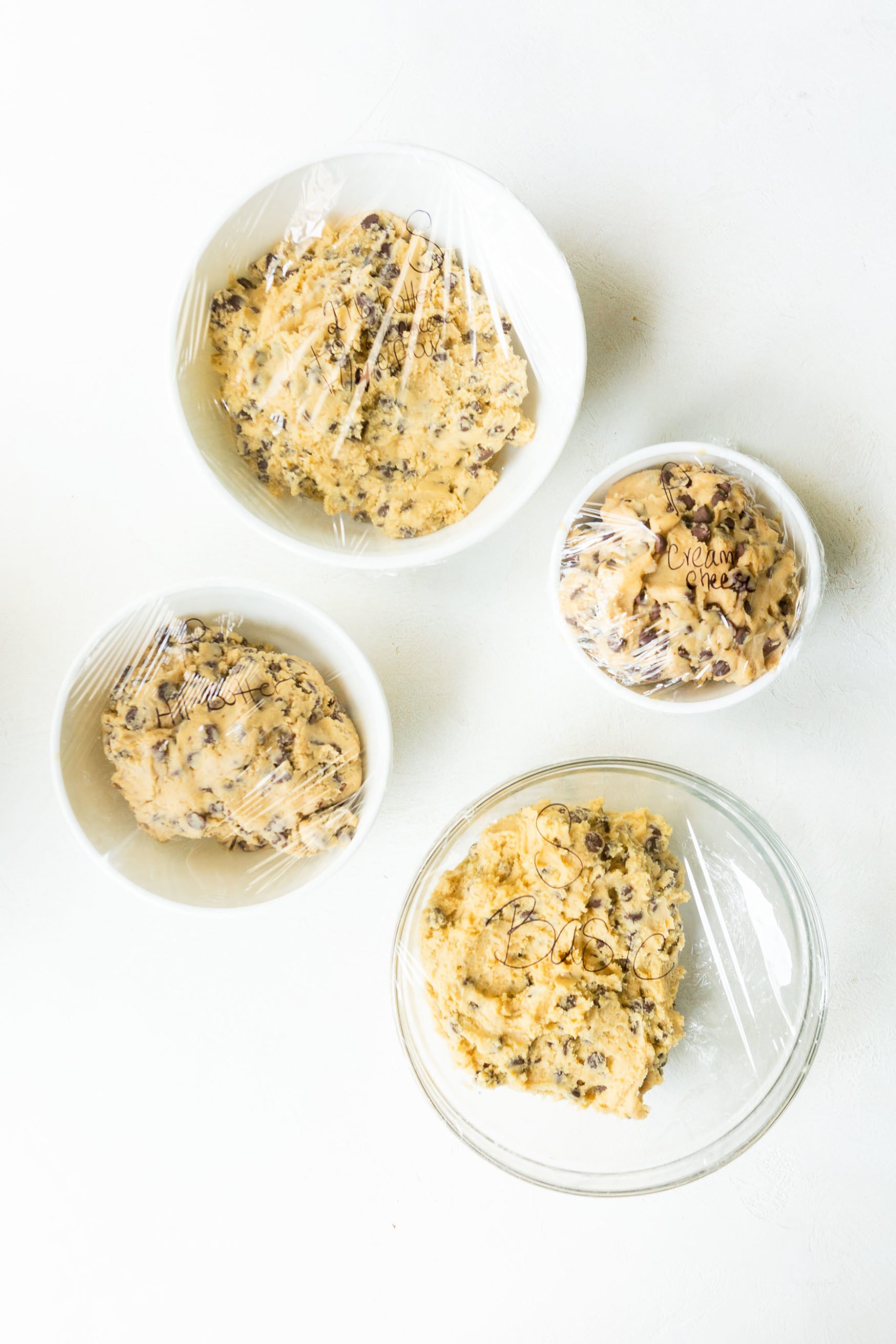Levain Bakery Style Thick Chocolate Chip Cookies Recipe a photo of four bowls of chocolate chip cookie dough sitting on a counter all with plastic wrap on them.