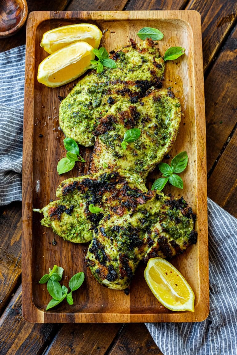 a photo of a large wooden serving board with grilled pesto chicken breasts and lime wedges sitting on it.