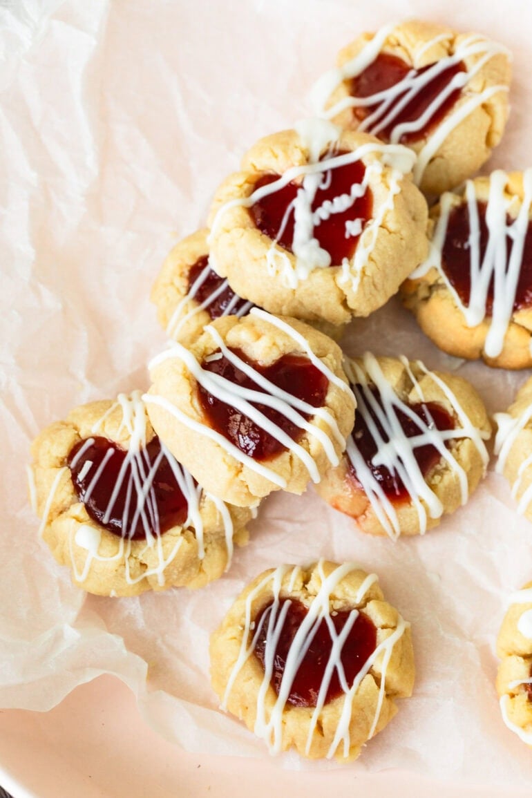 Overhead view of heart-shaped thumbprint cookies filled with red jam and drizzled with white chocolate.