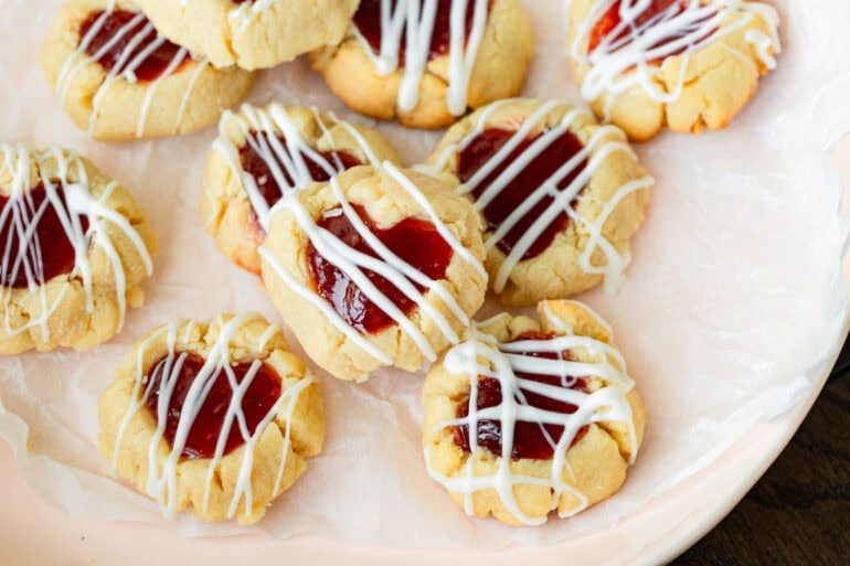 Valentine’s Day thumbprint cookies arranged on a plate with jam-filled centers and white chocolate drizzle.